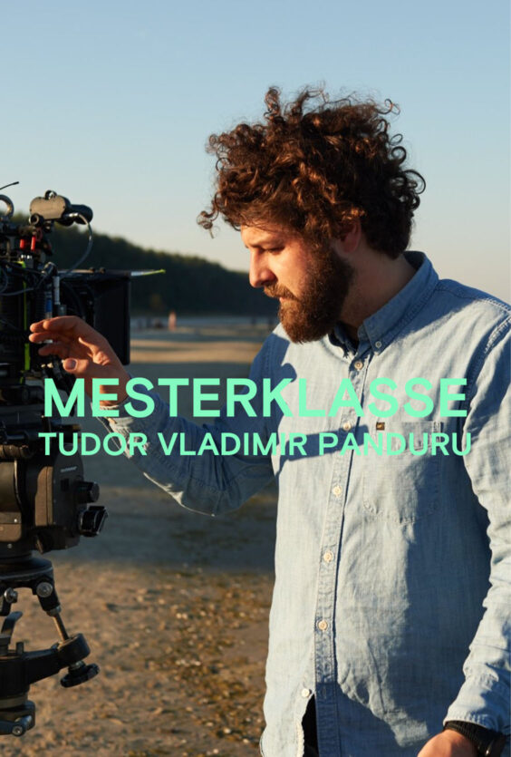 Mesterklasse. En mann med skjegg og krøllete hår i lyseblå skjorte står utendørs og justerer et stort filmkamera. Trær og strand i bakgrunnen.