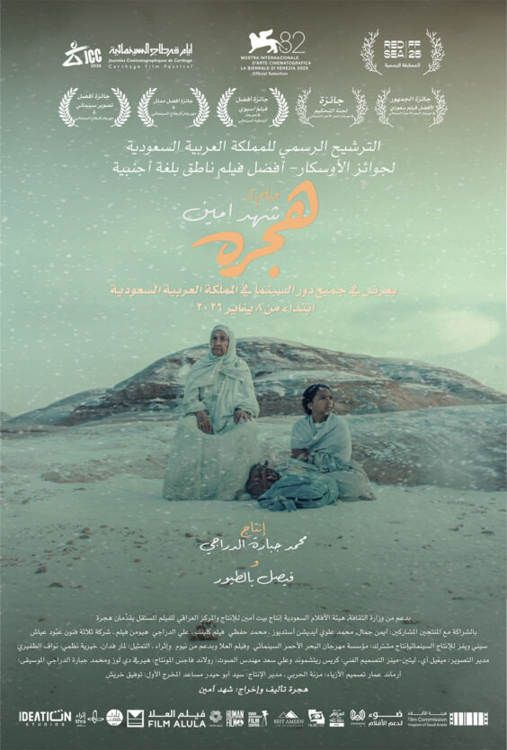 Hijra filmplakat med arabisk tekst og festivalmerker øverst og nederst To personer i lyse kapper sitter på sanden foran lave steinformasjoner