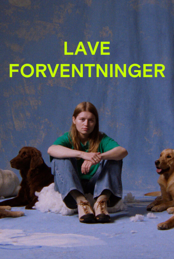 Lave Forventninger En kvinne med langt lyst hår sitter på gulvet foran et blått bakteppe To hunder sitter ved siden av henne