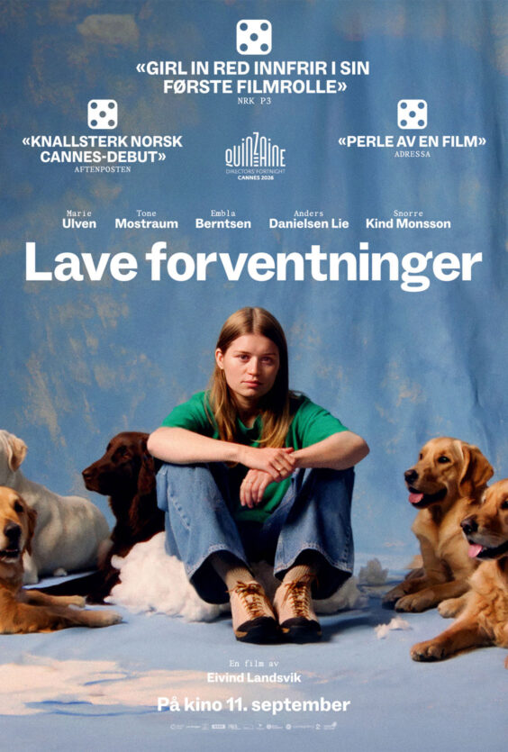 Lave Forventninger En kvinne med langt lyst hår sitter på gulvet foran et blått bakteppe To hunder sitter ved siden av henne