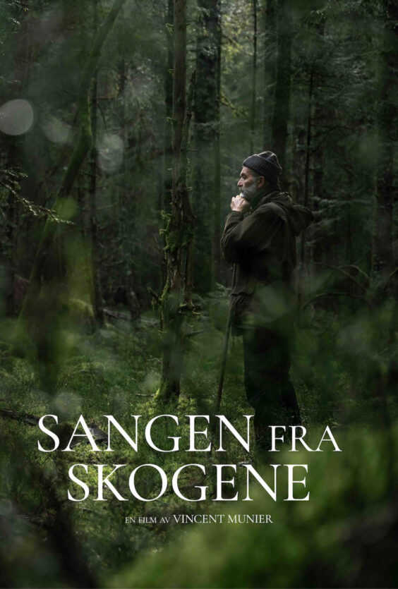 Sangen fra skogene En mann med skjegg og lue står i en grønn skog med trær og mose Han holder hendene mot ansiktet Han har på mørke klær