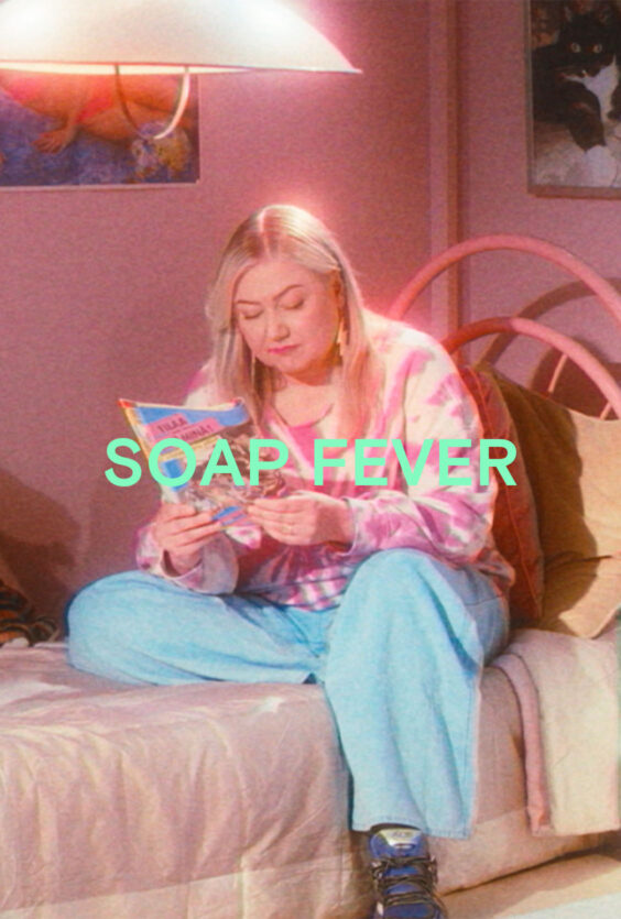 Soap Fever En kvinne med lyst hår sitter på en seng med rosa sengeteppe Hun har på seg rosa og hvit genser og lyseblå bukse Hun holder et blad Veggen bak er rosa